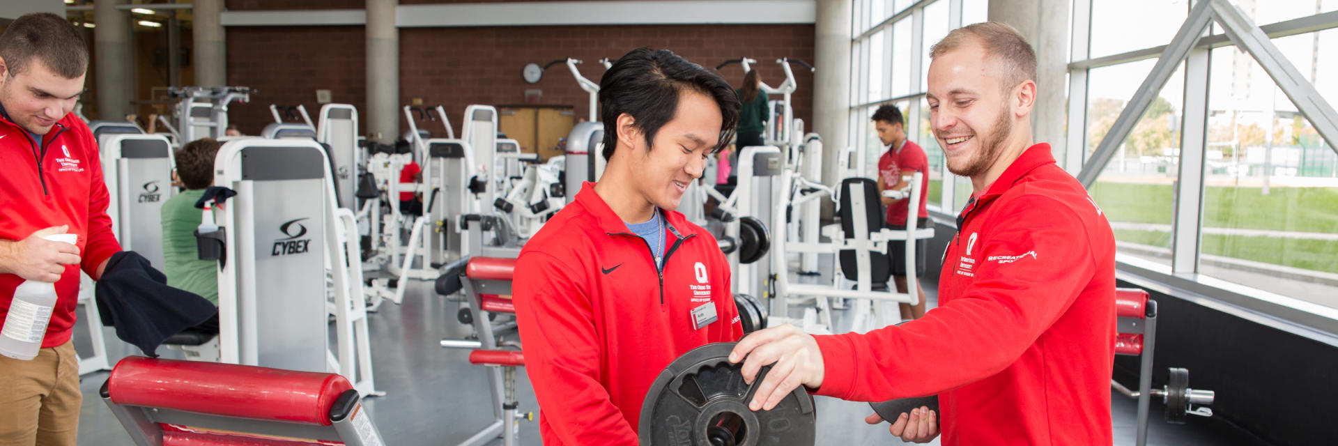 A handful of students in a gym are racking weights and cleaning equipment