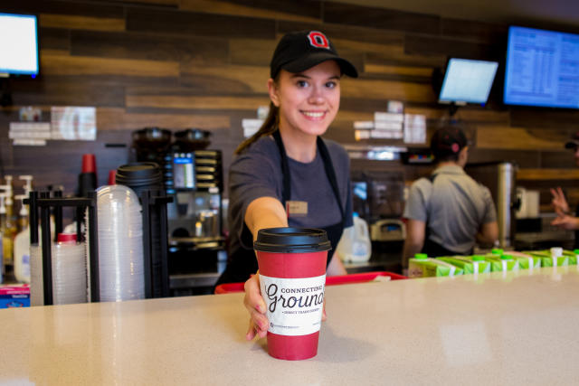 Smiling female student in background hands a cup of coffee toward the camera