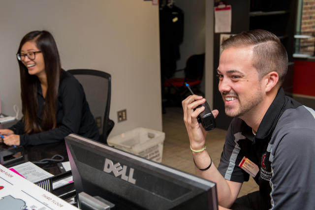 A male employee talks into a walkie talkie and a female students works at a computer